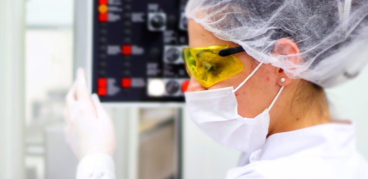 A woman wearing a lab coat and goggles is looking at a machine.