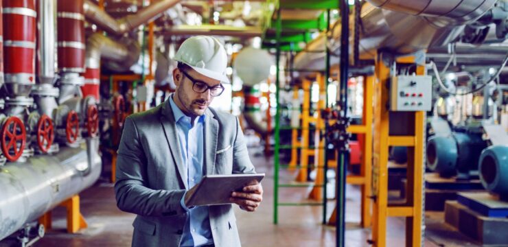 A man in a suit is using a tablet in a factory.