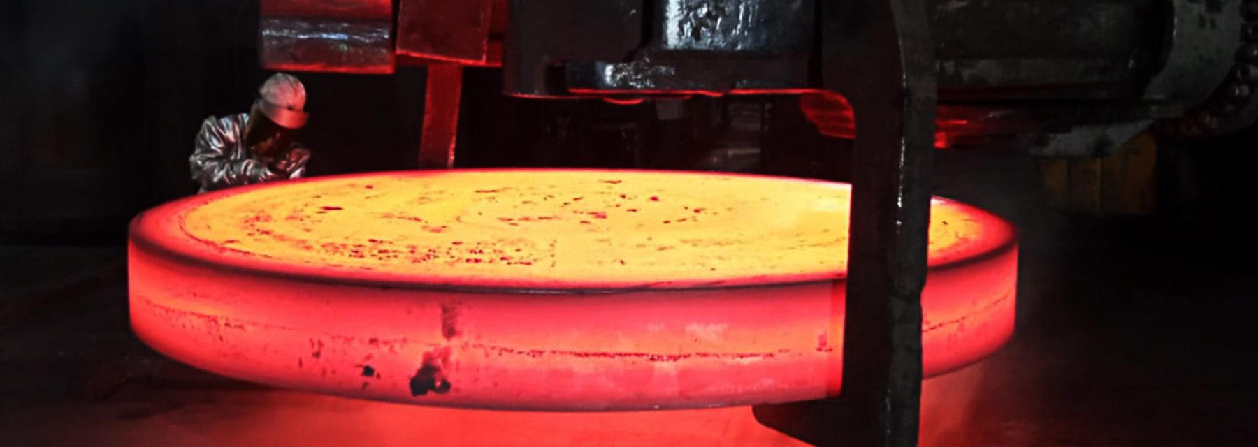 Worker inspecting a large, glowing red-hot metal disc in an industrial setting.