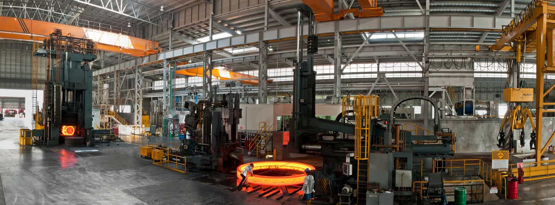 Panorama of an industrial steel foundry with machinery and large glowing hot metal ring, showing busy workflow under high ceiling.