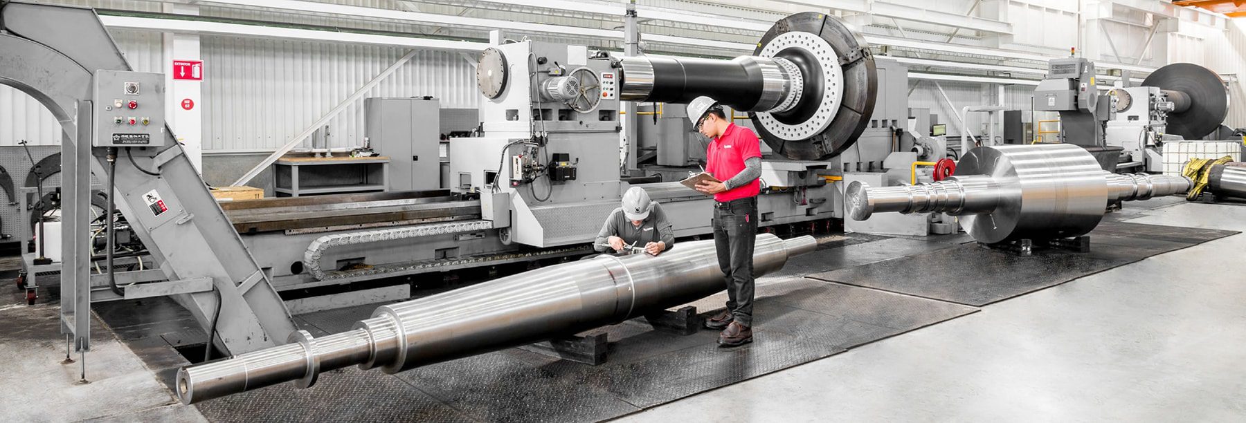 Worker inspecting a large metal shaft on a lathe machine in an industrial setting.